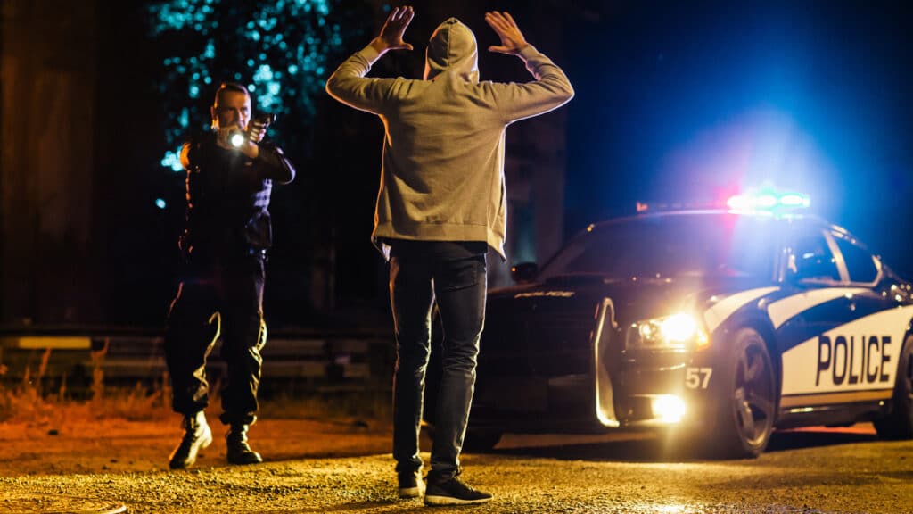 police officer and car pointing gun at hooded person with their hands up